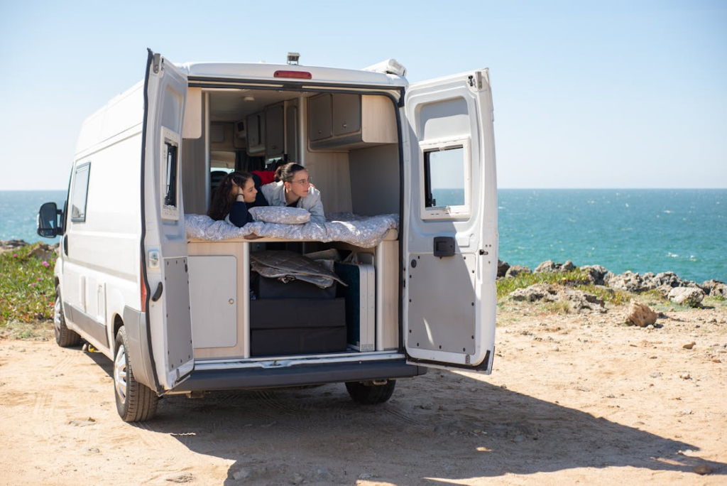 Two people lying in bed inside a campervan with its back doors open, looking out at the ocean.