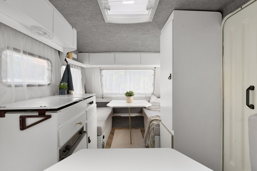 Interior view of a campervan with a white, clean design. Features include a kitchen counter, seating area with a table, and natural light coming through the windows.