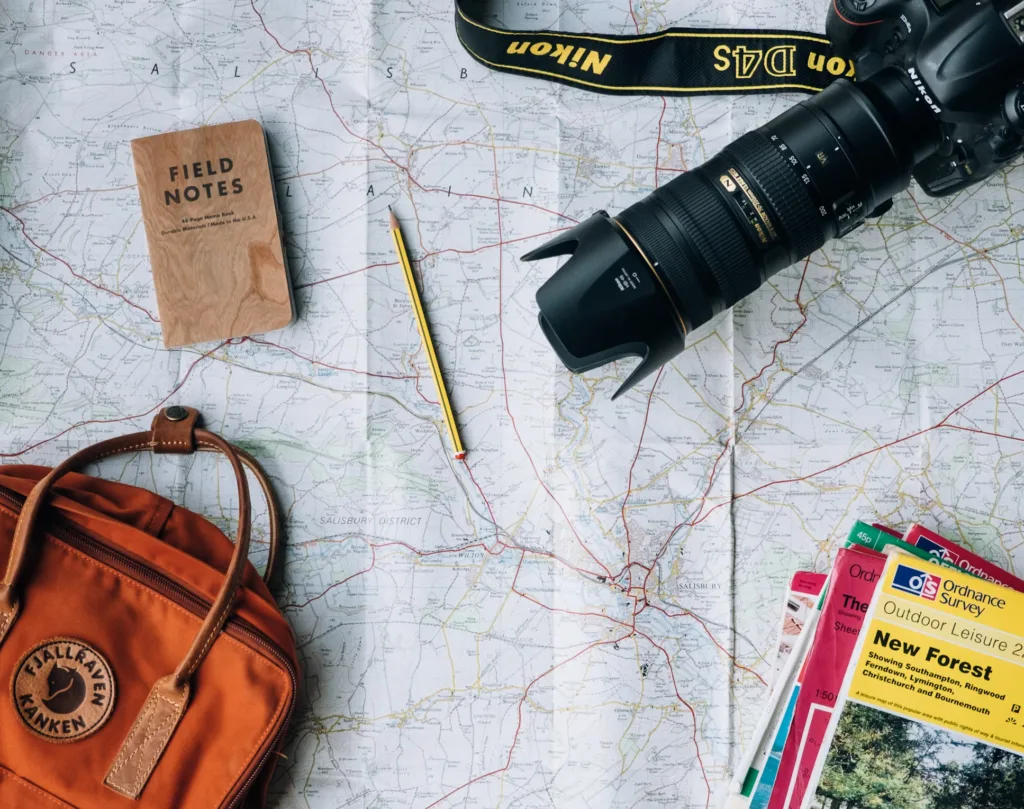 A flat lay image of a partially folded map spread out on a surface. On top of the map are various travel items: an orange backpack, a notebook labeled "Field Notes," a pencil, a DSLR camera with a telephoto lens, and several guidebooks and maps, including one prominently titled "New Forest."