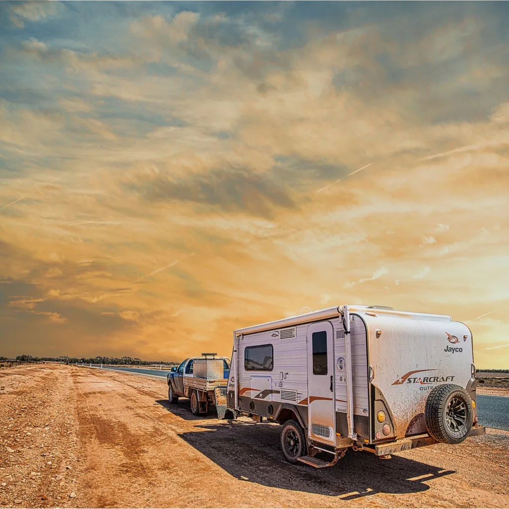 A blue pickup truck towing a small travel trailer on a dirt road at sunset.