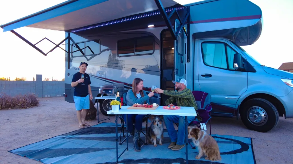 Three people and two dogs sitting and standing around a table set with food and drinks in front of a motorhome, with an awning extended