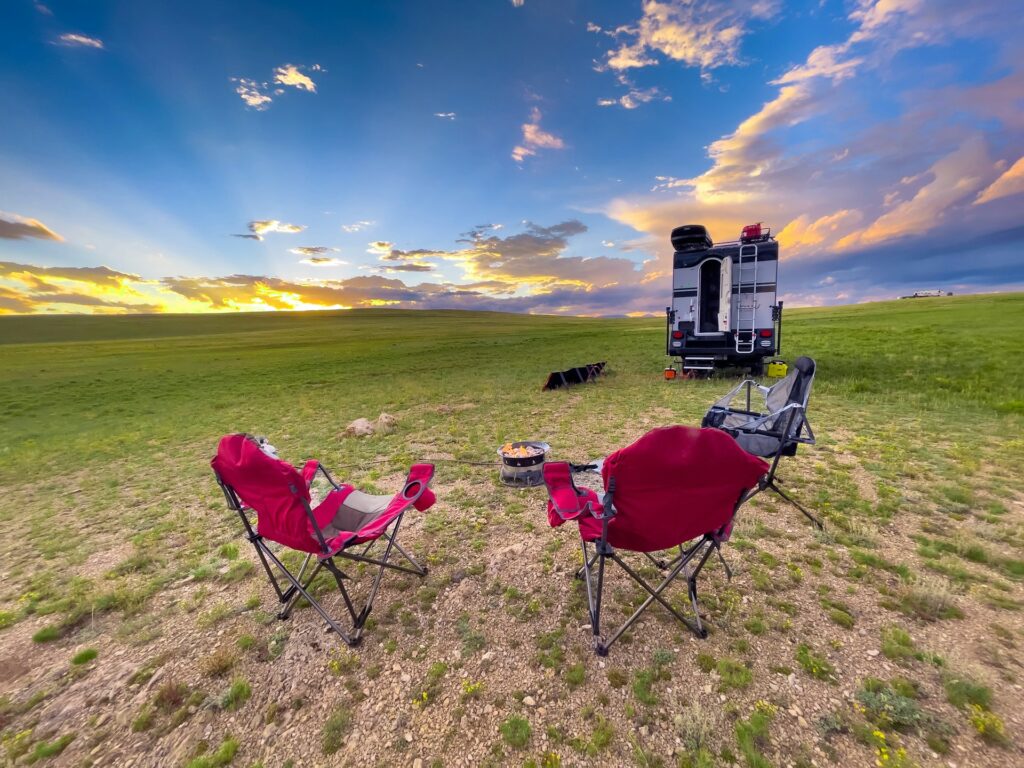 A picturesque campsite featuring a small RV parked in an open grassy field during sunset, with two empty camp chairs facing a campfire and the horizon.