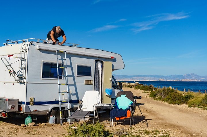 A man standing on the roof of a camper van adjusting equipment, with chairs and clothes drying nearby, next to a beach.