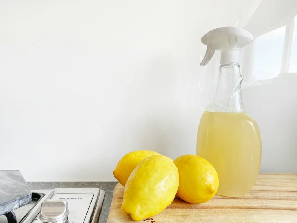 A glass spray bottle filled with lemon liquid sits next to three whole lemons on a wooden cutting board, against a white background.