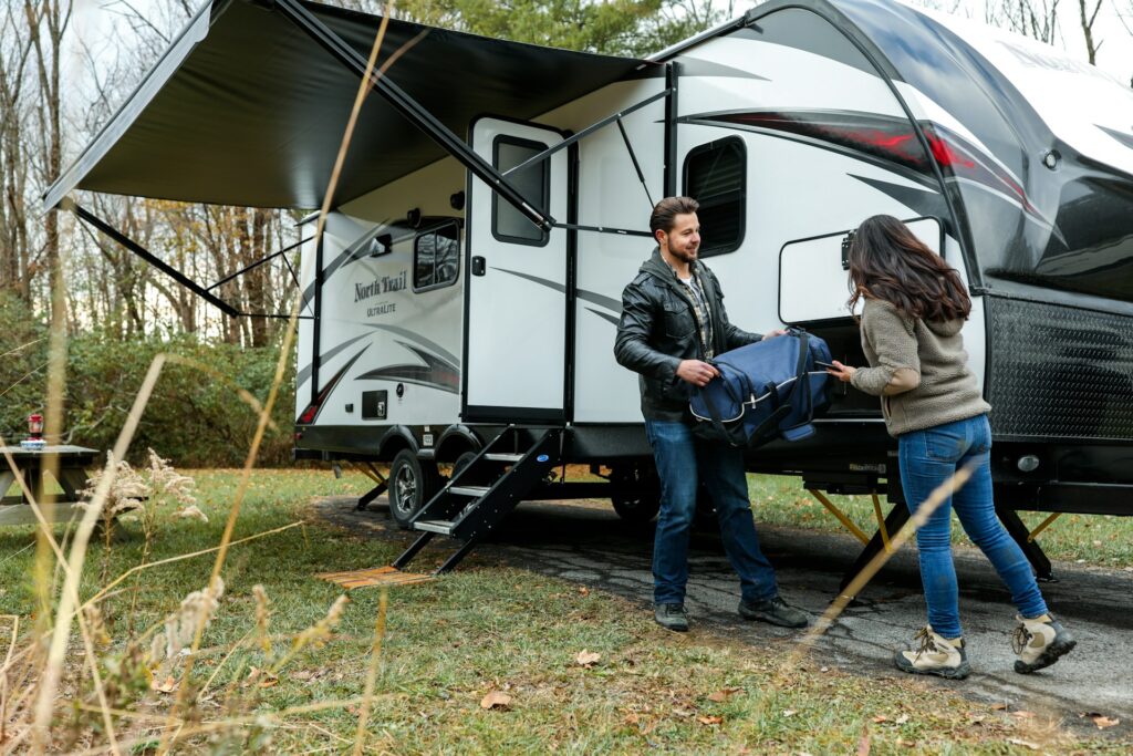 Two people unpacking or packing bags at a storage compartment of a motorhome parked in the woods.