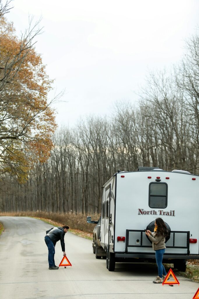 A person setting up an emergency warning triangle on the road while another person stands next to a "North Trail" camper. The camper is parked on the side of a forested road with bare trees on an overcast day.