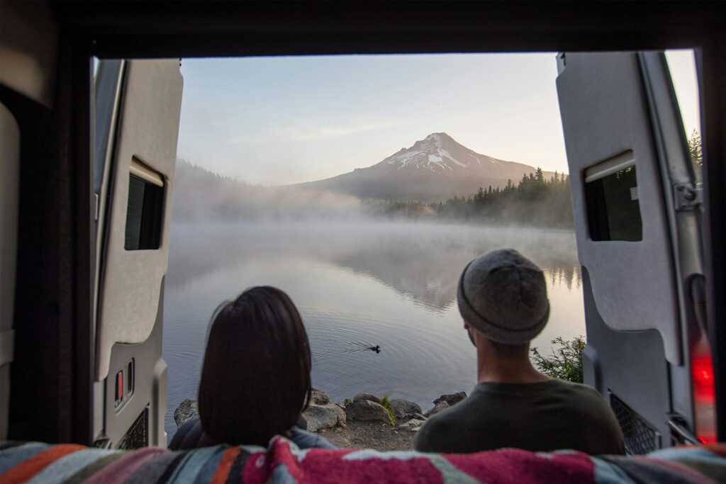 Two people sitting inside a camper van with the rear doors open, overlooking a serene lake with a snow-capped mountain in the background.