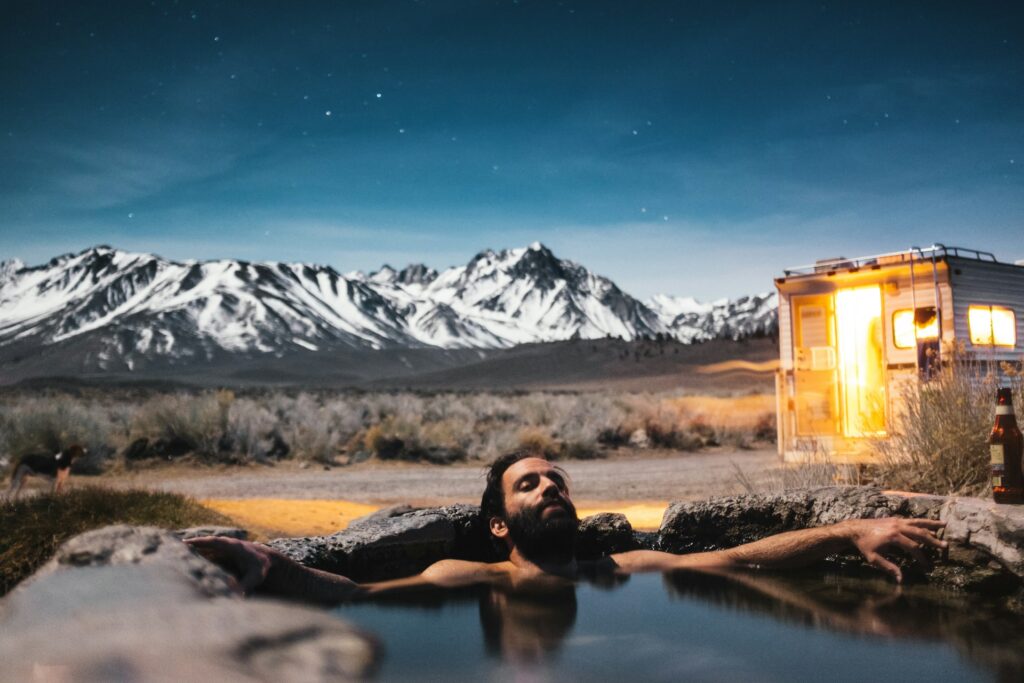 A man relaxing in a natural hot spring under a clear night sky with stars, surrounded by a landscape of snowy mountains. There is an illuminated camper van in the background and a beer bottle placed near the hot spring.