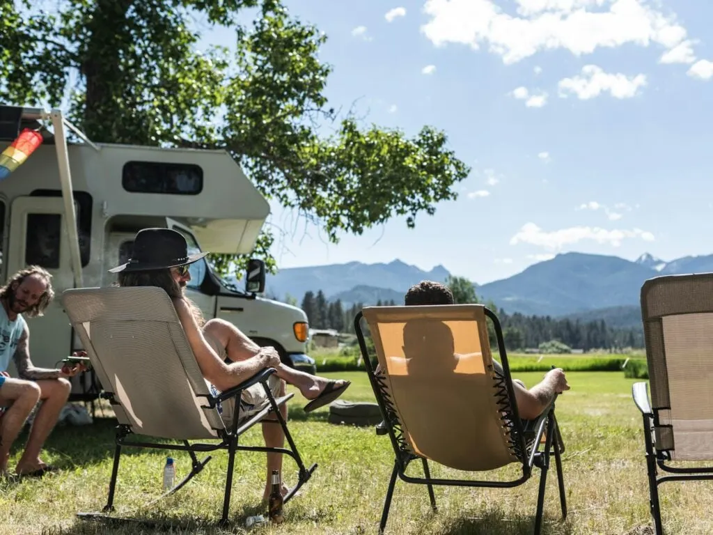 People relaxing in lawn chairs under a tree, with an RV and mountains in the background on a sunny day