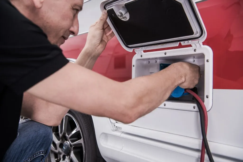 A person connecting an electrical plug to an outlet on the side of a white and red camper van.