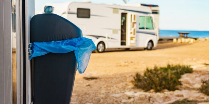 A close-up of a trash bin with a blue bag, parked near a motorhome with the sea in the background.