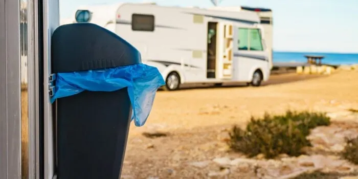 A close-up of a trash bin with a blue bag, parked near a motorhome with the sea in the background.