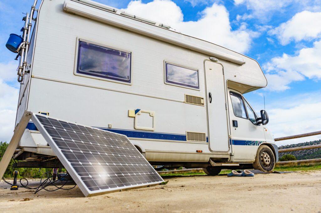 A white and blue motorhome parked outdoors under a clear, blue sky, with a solar panel set up on the ground beside it.