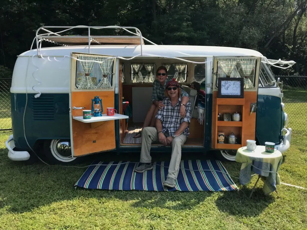 Two people sitting together inside the open doorway of a vintage camper van, which is set up with a small table, mugs, and additional decorations on a grassy area.