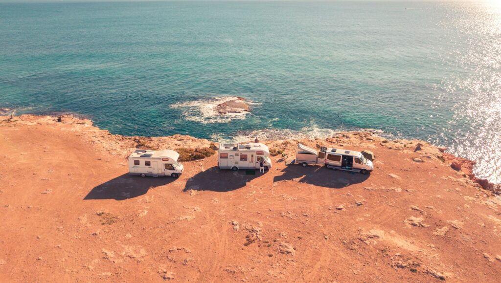 Three campervans parked on a rocky cliff overlooking the ocean, with sunlight reflecting off the water.