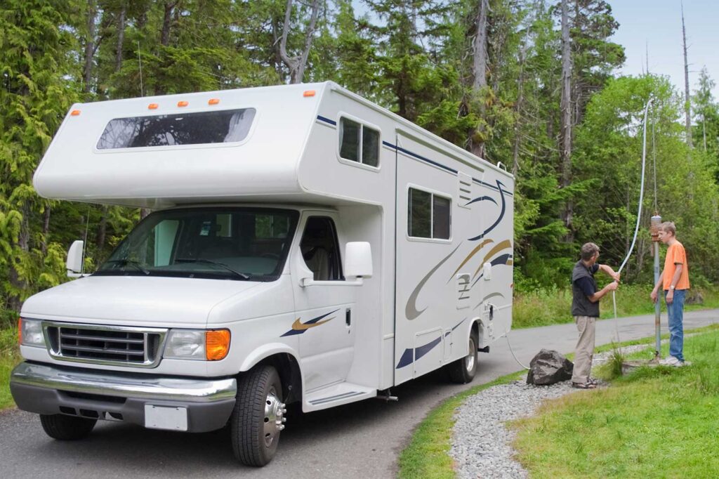 A motorhome parked on a campsite driveway surrounded by trees, with two people working near a utility pole.