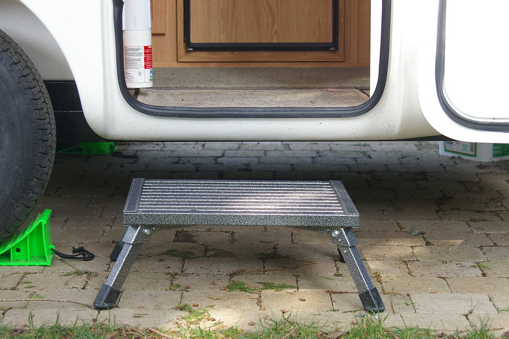 A small metal step stool placed outside the open door of a white camper van, making it easier to step into the vehicle. The van is parked on a paved surface, equipped with green leveling blocks beneath the wheels. The door offers a glimpse of the wooden interior and what appears to be a fire extinguisher.