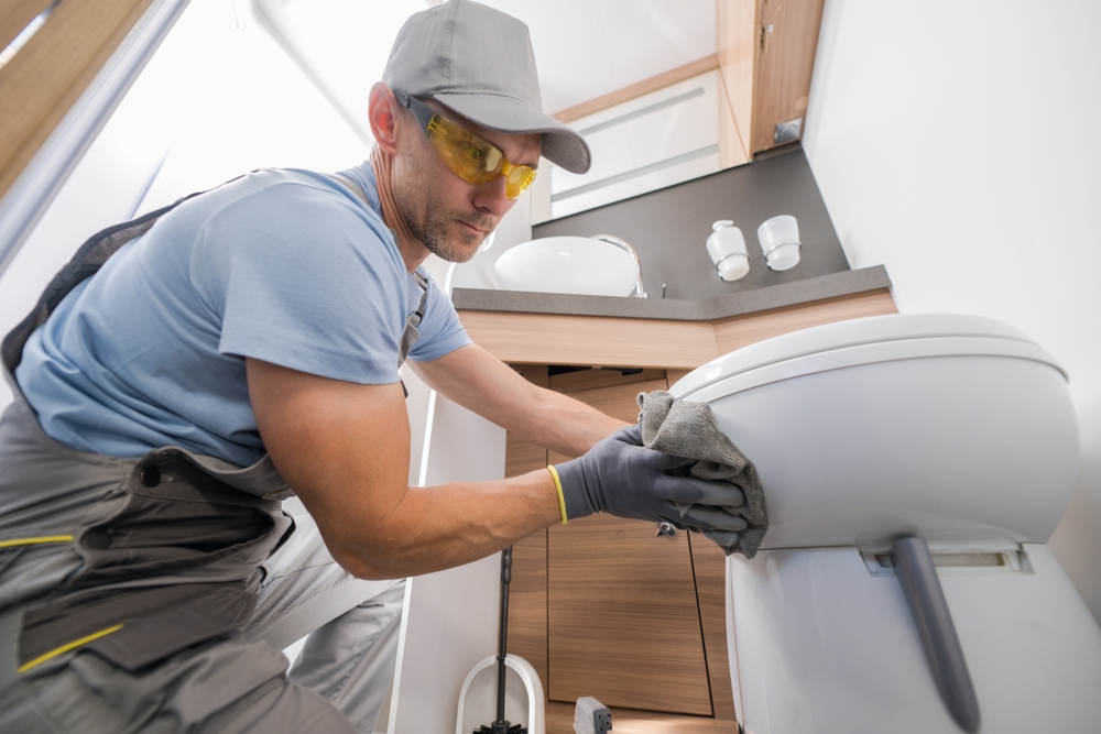 A person in work attire, wearing gloves and glasses, cleaning a toilet inside an RV or camper bathroom with a cloth.