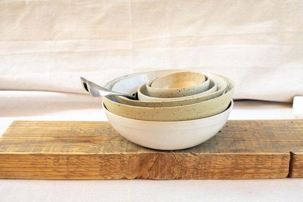 A set of nested ceramic bowls of varying sizes, stacked neatly inside each other, placed on a rustic wooden board. A silver spoon rests in one of the bowls. The background features a neutral-colored fabric backdrop.