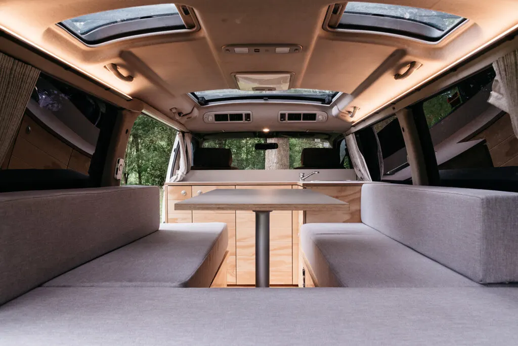 The interior of a camper van featuring a central table surrounded by cushioned benches, skylights, and wooden cabinetry at the rear.