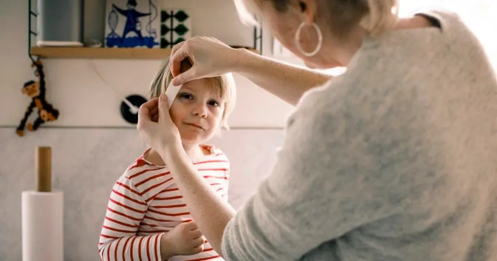 A woman applying a bandage to a child's forehead, with the child looking ahead and wearing a striped shirt in a cozy indoor setting.