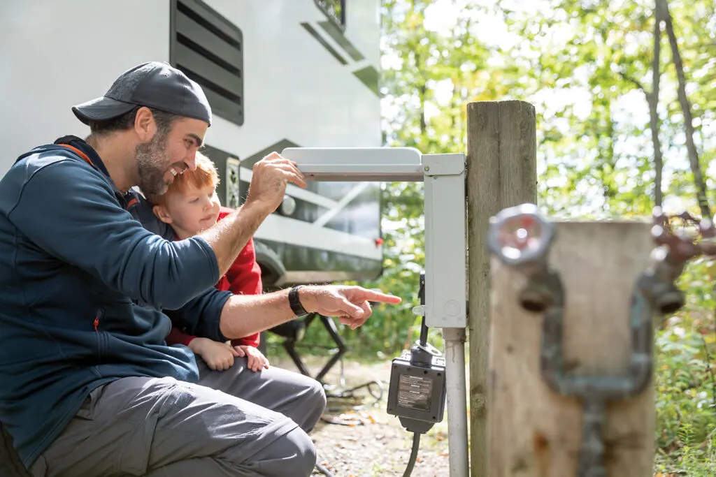 A man wearing a baseball cap and a young boy are examining an outdoor electrical hookup next to their camper van. The man is opening the cover while the boy watches closely. The background has lush greenery and the camper van is partially visible.