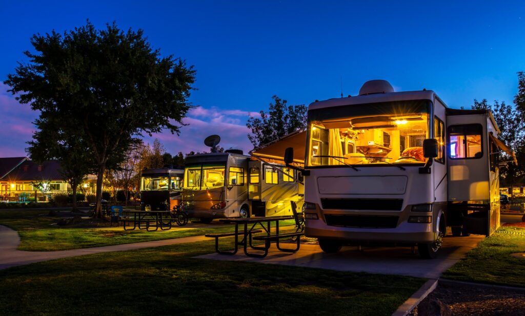 Several RVs parked at a campsite during the evening, with warm interior lights glowing, picnic tables, and surrounding trees against a twilight sky.