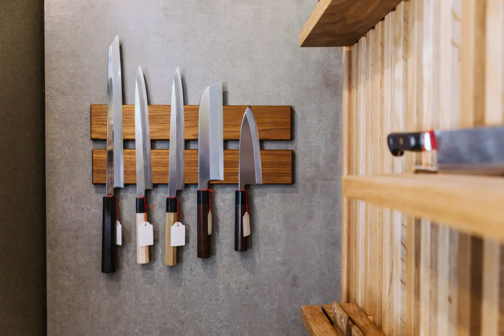 A display of six sharp kitchen knives with wooden handles hanging on a wooden magnetic knife rack mounted on a concrete wall. The knives vary in size and shape, and each has a small tag attached to the handle. The wall is next to wooden shelves that hold additional kitchen tools.