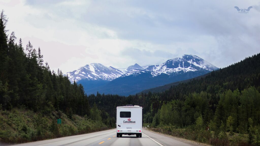 An RV driving on an open road surrounded by dense forest, with snow-capped mountains visible in the distance under a cloudy sky.