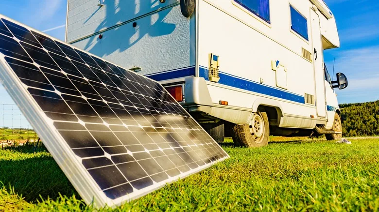 A camper van parked on a grassy field, with a large solar panel propped up in front of it, angled towards the sun. The sunlight reflects off the solar panel, highlighting its grid-like surface. The camper van has blue accents and is positioned to take advantage of the clear, sunny day.