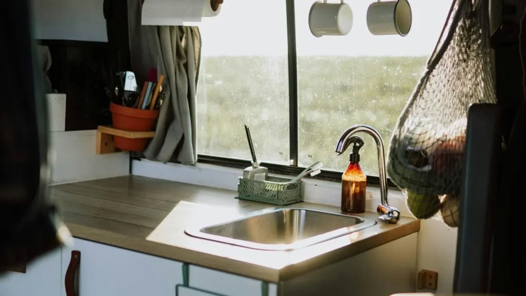  A compact kitchen area in the RV with a sink, faucet, utensils in a pot, hanging cups, and netted produce bag, with sunlight streaming through a window.