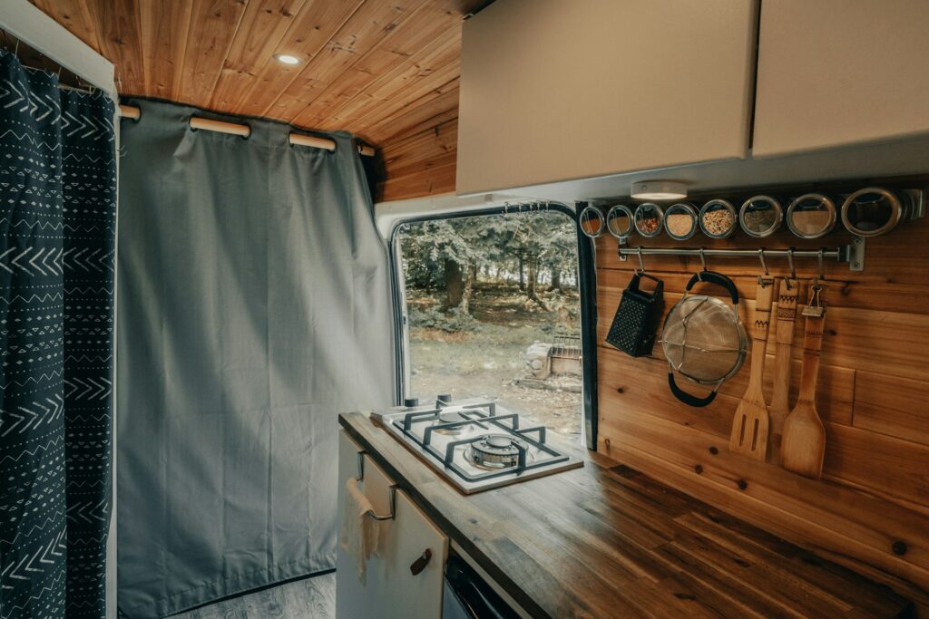 An interior view of a camper van kitchen with wooden cabinets, a gas stove, utensils hanging on the wall, and a curtain.