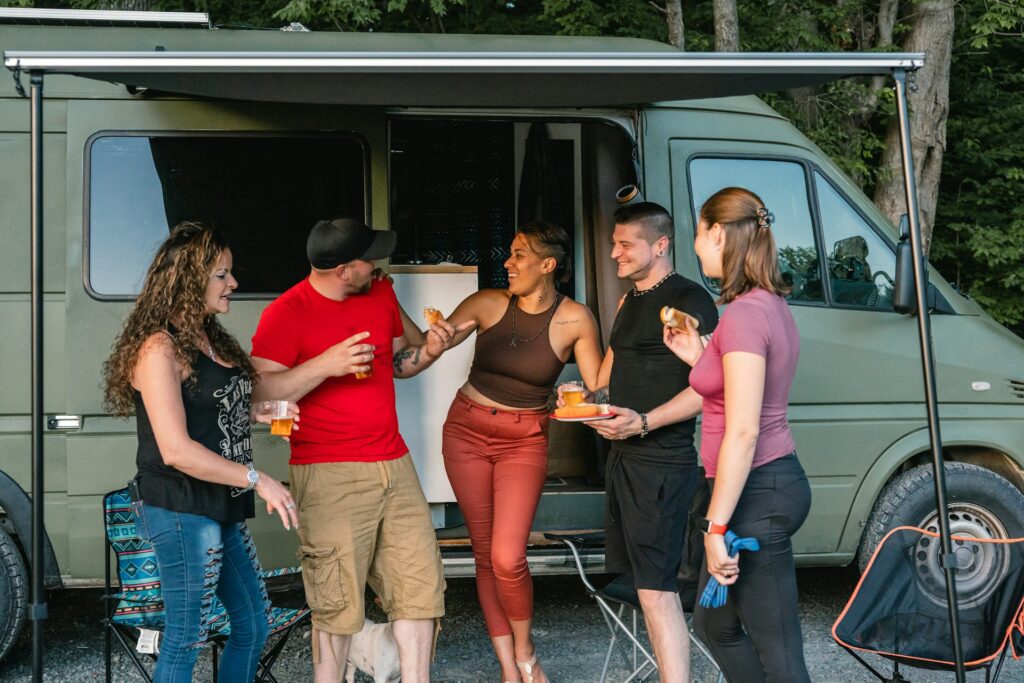 A group of friends standing and chatting outside a camper van, enjoying drinks and food with a relaxed and happy atmosphere. The awning of the camper van is extended, providing some shade, and the surrounding area is wooded.