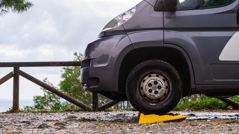 A close-up side view of a parked gray van with its front tire resting on a yellow wheel chock on a wet, gravelly surface. The background includes a wooden fence, greenery, and partly cloudy skies.