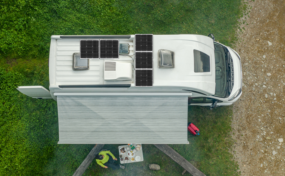 An aerial view of a camper van parked on a grassy area, equipped with solar panels on its roof. An awning extends out from the side, providing shade, and a person is seen sitting at a table under the awning, preparing food. The background includes grass and a gravel pathway.