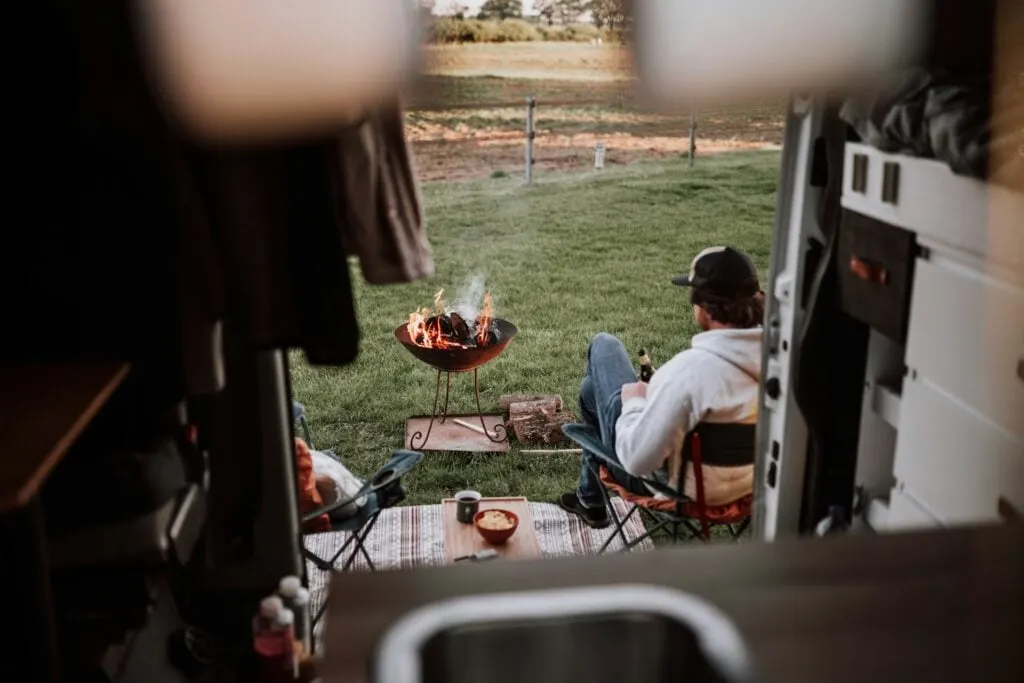 A view from inside a camper van looking out at a peaceful campsite. A person is sitting in a foldable chair, relaxing and holding a beverage, while a fire burns in a portable fire pit on the grass. There is a small table with snacks and drinks beside the person, and beyond the campsite, there is an open, grassy field with trees in the background. The scene exudes a tranquil, outdoor atmosphere.