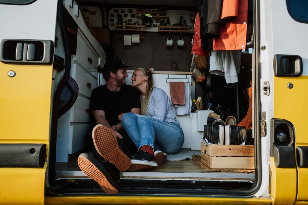 A couple sitting inside the open back door of a yellow and white camper van, smiling and looking at each other. The interior is cozy and organized, featuring a small kitchen area with cabinets, hanging mugs, and various personal items. The couple is casually dressed and appears happy and relaxed.