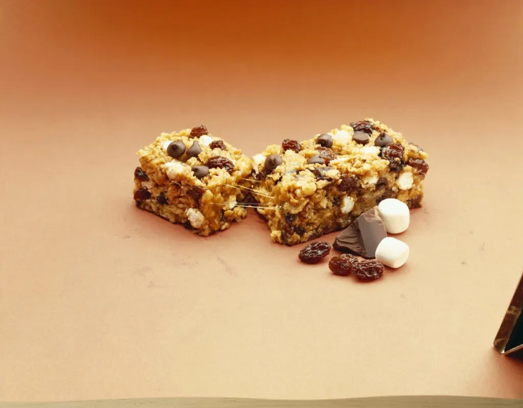 Two granola bars with chocolate chips, raisins, and miniature marshmallows, placed on a light brown surface. Nearby, there are additional raisins, chocolate pieces, and two small marshmallows.