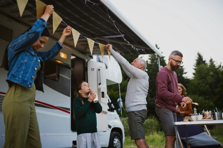 A group of people setting up decorations at a campsite. two individuals are hanging triangular bunting under the awning of a camper van, while a young girl stands nearby, eating and looking up. Another person is arranging items on a table set with food and drinks. Trees can be seen in the background, creating a pleasant outdoor setting.