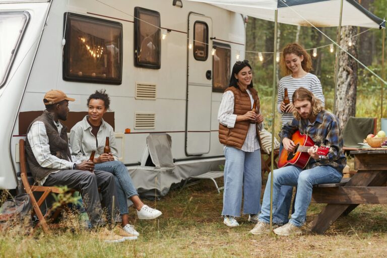 A group of five people enjoying time outside a caravan set up with string lights. Two people sit and converse with drinks, while another plays a guitar. Two others stand nearby, smiling and holding bottles. They are gathered around a picnic table with food and drinks.