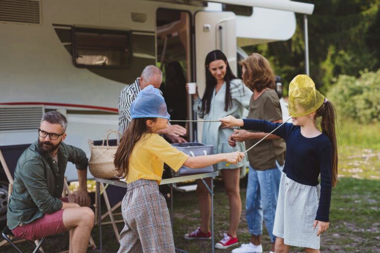 A group of people gathered near a camper van parked in a grassy area. Two children in the foreground are playing with netting on their heads, while adults in the background engage in conversation and prepare food or drinks.