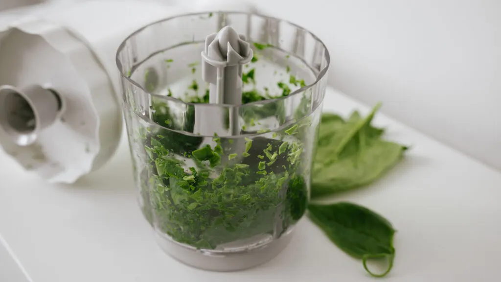 A close-up of a food processor containing freshly chopped green herbs, with some leaves scattered on the white surface beside it. The lid of the food processor is removed and placed next to the container.