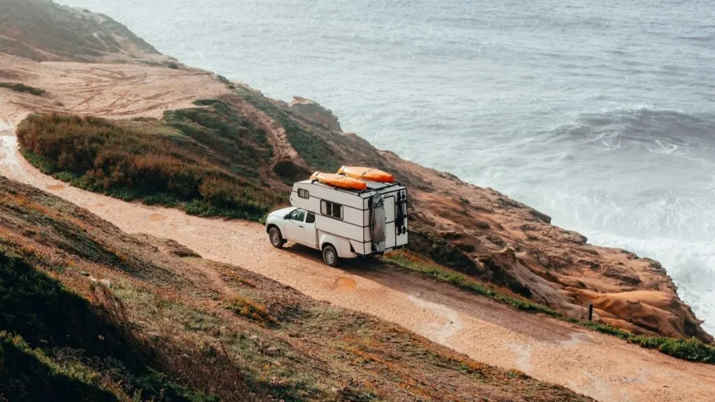 A camper truck parked on a rugged, sandy coastal trail with two orange kayaks secured on the roof. The scene overlooks a dramatic cliffside with the ocean waves crashing against the shore, surrounded by lush greenery and patches of vegetation on the hillside.