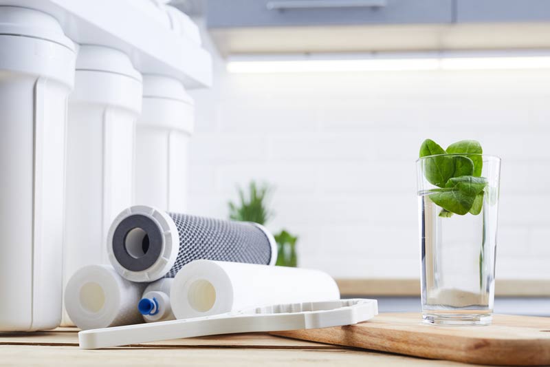 A set of water filter cartridges and components arranged on a wooden surface in a bright kitchen. Next to the filters is a clear glass of water with fresh green leaves inside. The background includes a white tiled backsplash and a blurred view of kitchen cabinets.