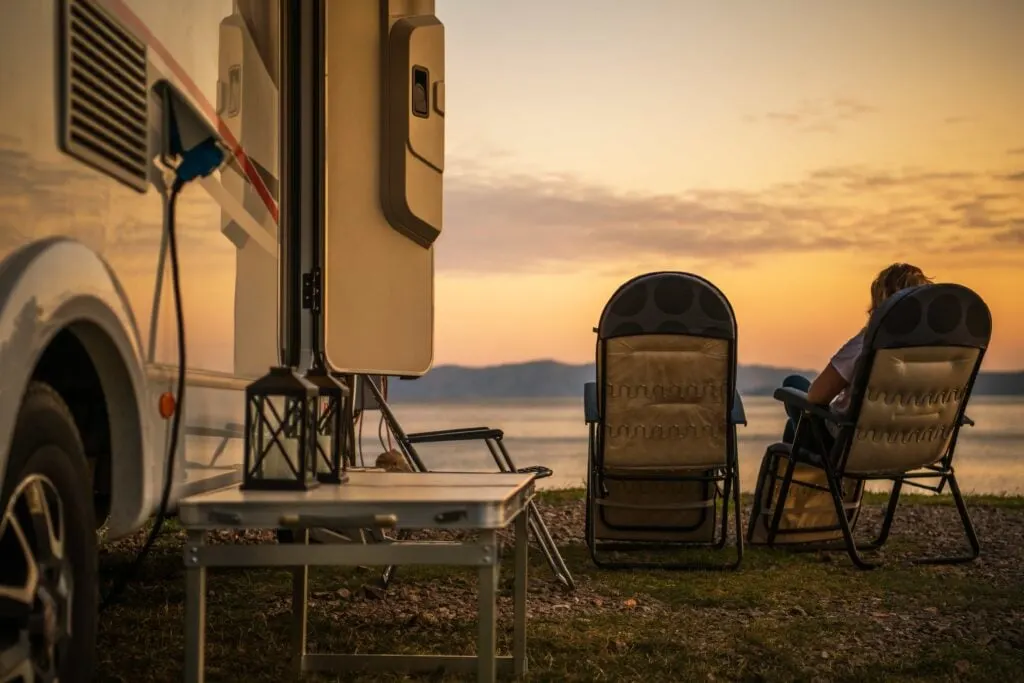 A serene scene at sunset, featuring a camper van parked near a body of water. Two comfortable outdoor chairs are set up facing the sunset, with a person relaxing in one of them. A small foldable table beside the van holds two lanterns. The sky is beautifully illuminated with warm hues of orange and yellow as the sun sets over the distant hills and water, creating a peaceful and tranquil atmosphere.