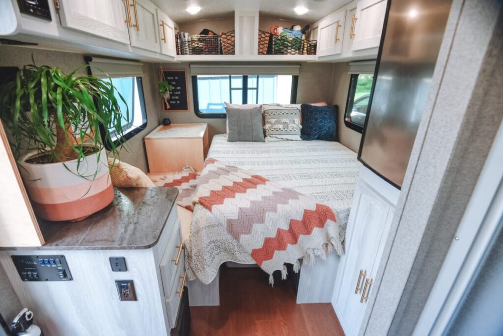 A cozy and well-decorated interior of a camper van bedroom featuring a neatly made bed with patterned bedding and throw pillows. There are storage cabinets above and around the bed, a small side table with a potted plant, and a woven blanket draped on the bed. The room is illuminated by natural light from the windows.