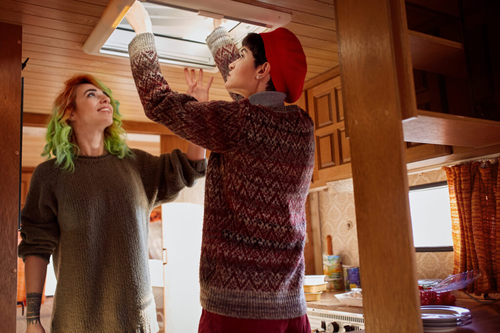 Two people working inside a camper, one adjusting the skylight while the other looks on with a smile, in a cozy wooden-paneled interior.