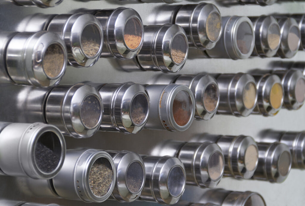 A collection of stainless steel spice containers with transparent lids mounted on a wall or magnetic surface. Each container holds different spices and seasonings, visible through the lids. The containers are neatly organized in rows and columns.