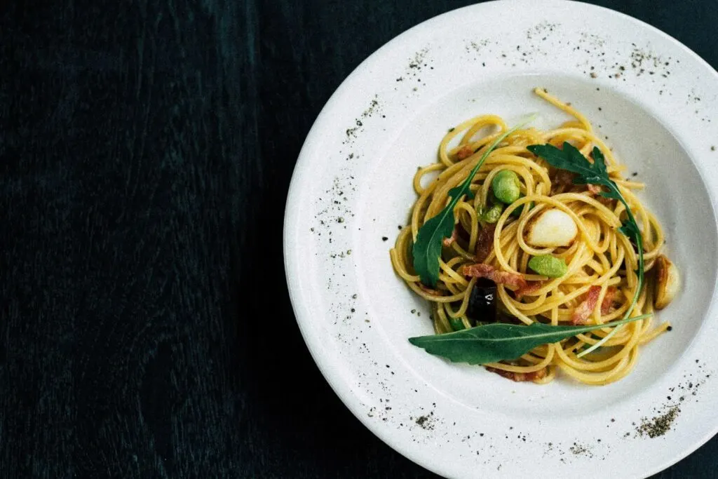 A plate of spaghetti with vegetables and arugula, sprinkled with black pepper, on a dark background.