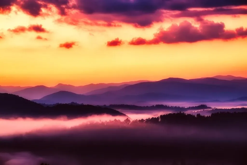 A breathtaking sunset view of layered, mist-covered mountain ranges in shades of purple and blue in Great Smoky Mountains National Park. The sky is ablaze with vibrant colors of orange, pink, and yellow, with some clouds adding depth to the scene.